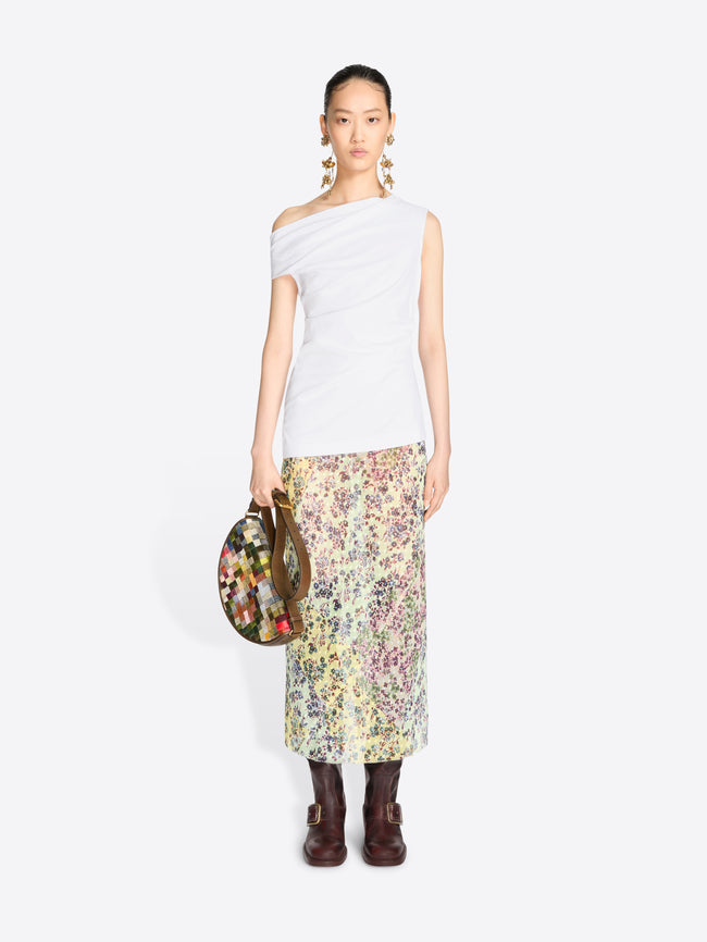A model stands against a light gray background wearing a white asymmetrical off-the-shoulder top and a long floral print midi skirt, holding a round floral handbag.