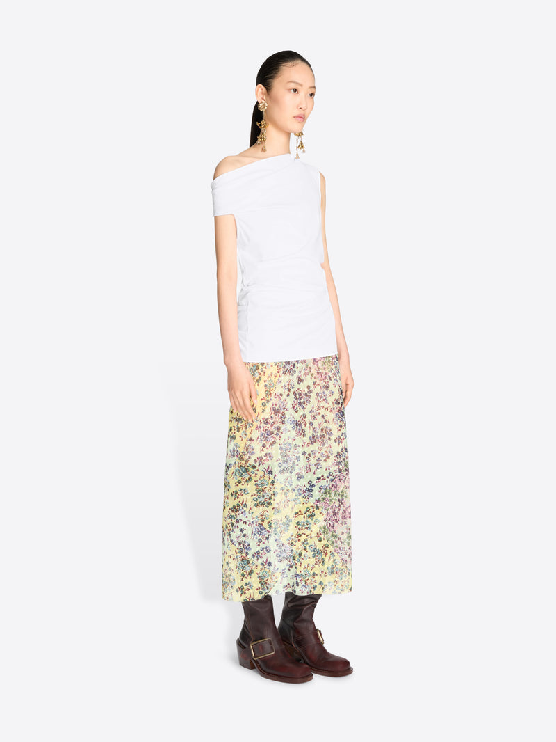 A model stands against a light gray background wearing a white asymmetrical off-the-shoulder top and a long floral print midi skirt.