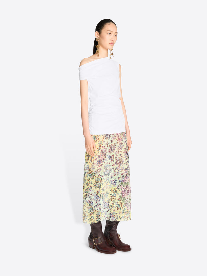 A model stands against a light gray background wearing a white asymmetrical off-the-shoulder top and a long floral print midi skirt.