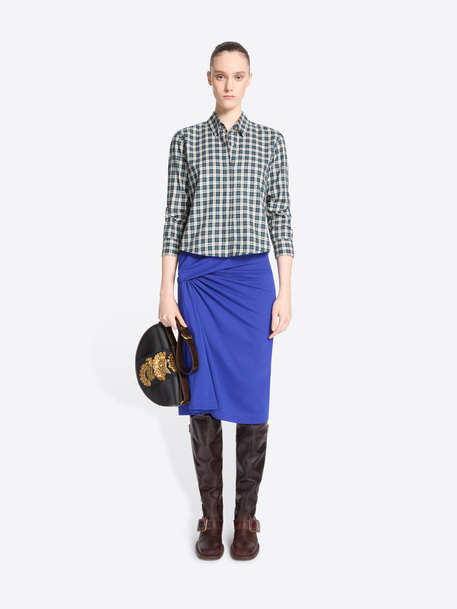 A model stands against a light gray background wearing a green and white checkered button-up shirt, a bright royal blue wrap skirt, dark brown leather boots, and holding a black handbag with a gold detail.