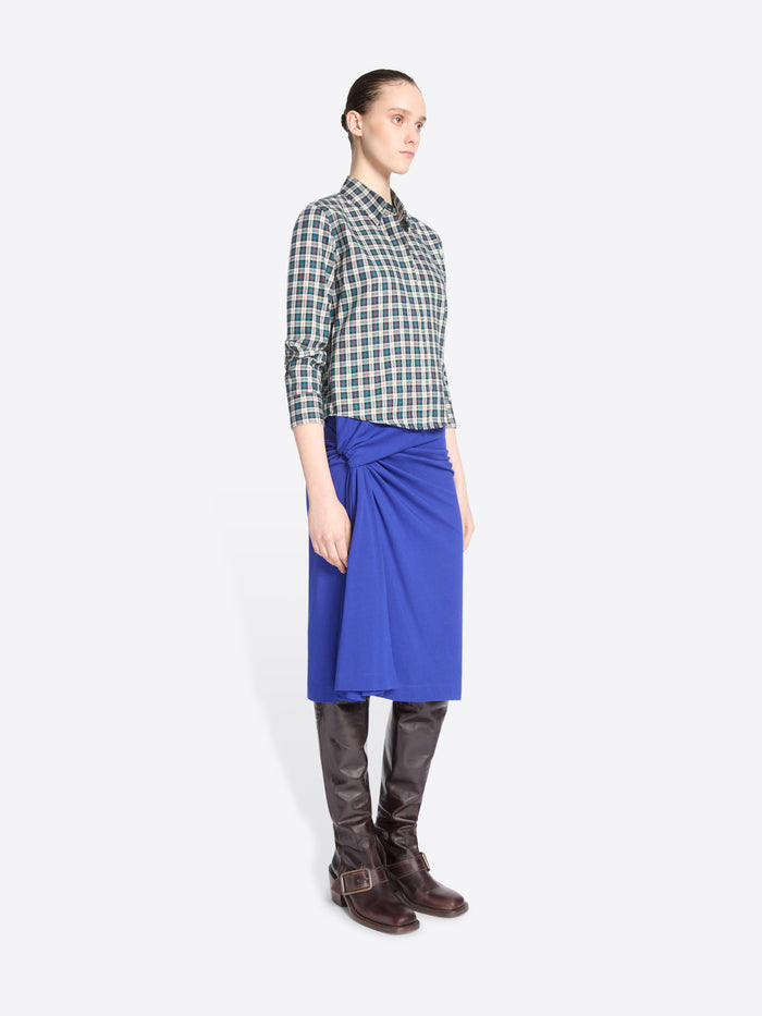 A model stands against a light gray background wearing a green and white checkered button-up shirt, a bright royal blue wrap skirt, dark brown leather boots.