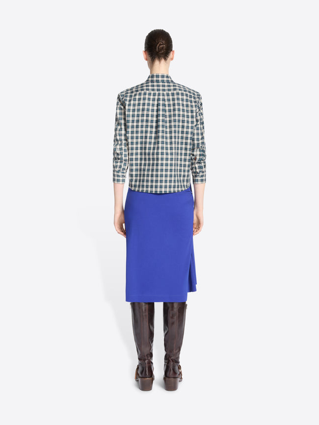 Back view of a model standing against a light gray background wearing a green and white checkered button-up shirt, a bright royal blue wrap skirt, dark brown leather boots.