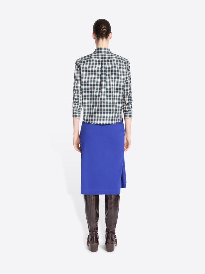 Back view of a model standing against a light gray background wearing a green and white checkered button-up shirt, a bright royal blue wrap skirt, dark brown leather boots.