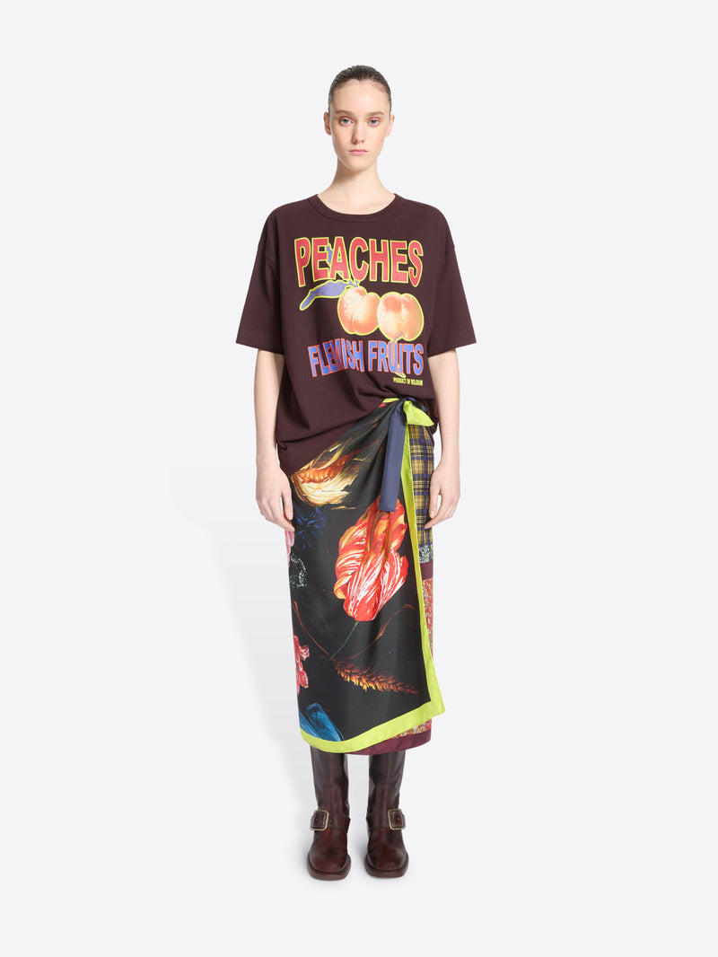 A model stands against a light gray background wearing a dark brown graphic T-shirt with peach imagery and the words 'PEACHES FLEMISH FRUITS,' paired with a long black wrap skirt featuring a vivid floral print and neon green trim and dark brown ankle boots.