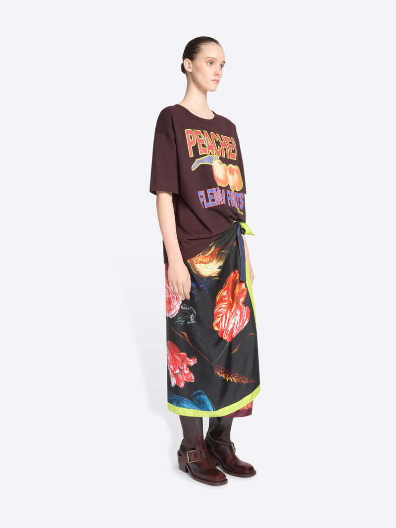 A model stands against a light gray background wearing a dark brown graphic T-shirt with peach imagery and the words 'PEACHES FLEMISH FRUITS,' paired with a long black wrap skirt featuring a vivid floral print and neon green trim and dark brown ankle boots.