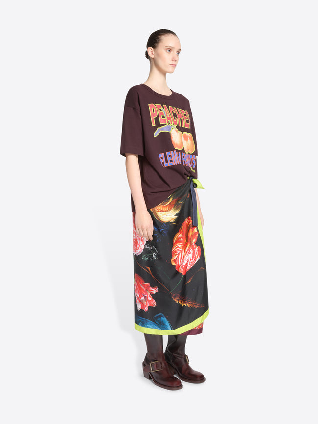 A model stands against a light gray background wearing a dark brown graphic T-shirt with peach imagery and the words 'PEACHES FLEMISH FRUITS,' paired with a long black wrap skirt featuring a vivid floral print and neon green trim and dark brown ankle boots.