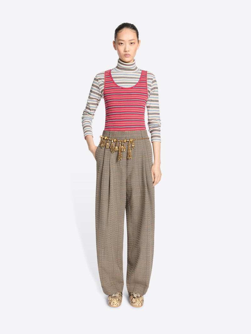 A model stands against a light gray background wearing a striped turtleneck top, a striped tank top, wide-leg pleated pants, and snakeskin shoes.