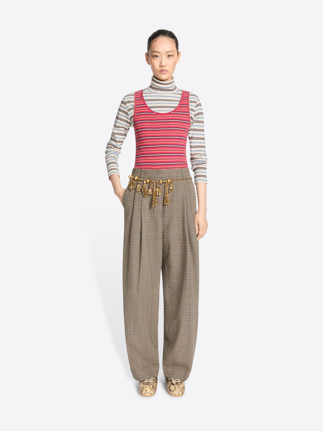 A model stands against a light gray background wearing a striped turtleneck top, a striped tank top, wide-leg pleated pants, and snakeskin shoes.