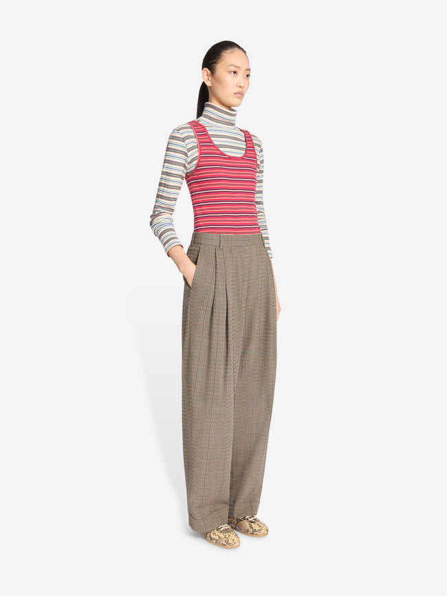 A model stands against a light gray background wearing a striped turtleneck top, a striped tank top, wide-leg pleated pants, and snakeskin shoes.