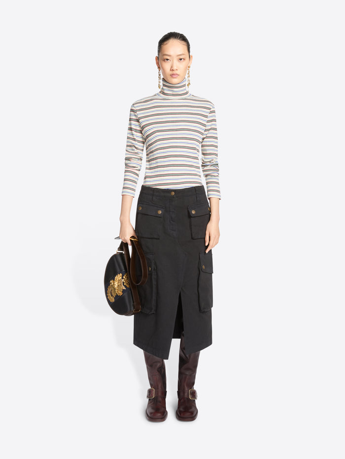 A model stands against a light gray background wearing a striped turtleneck sweater, a black cargo midi skirt with front flap pockets, dark brown knee-high boots, and holding a black round handbag with gold embroidery.