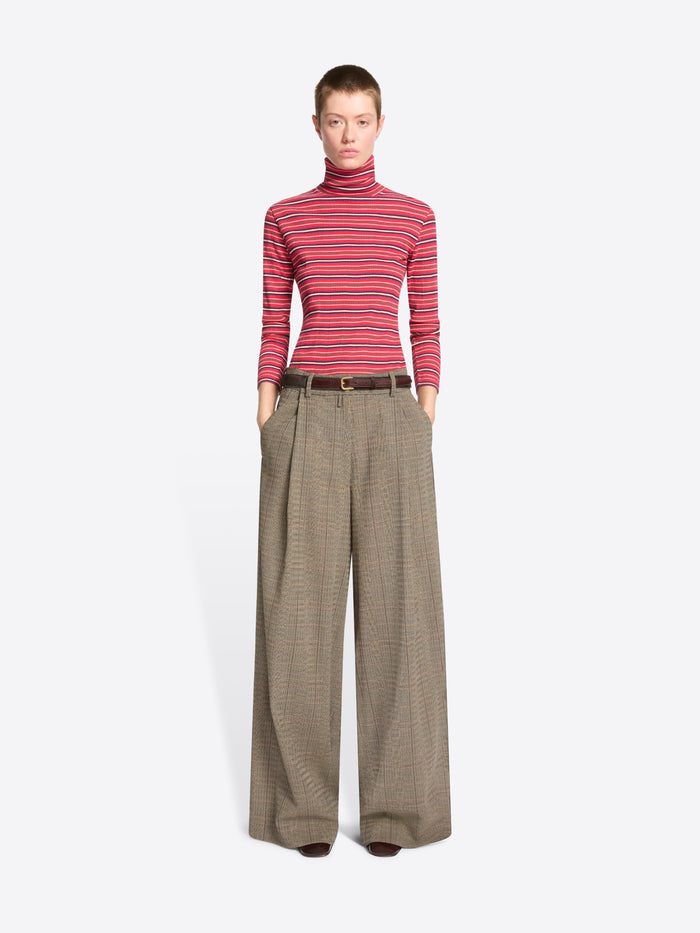 A model stands against a light gray background wearing a red and pink striped turtleneck sweater paired with wide-leg brown trousers.
