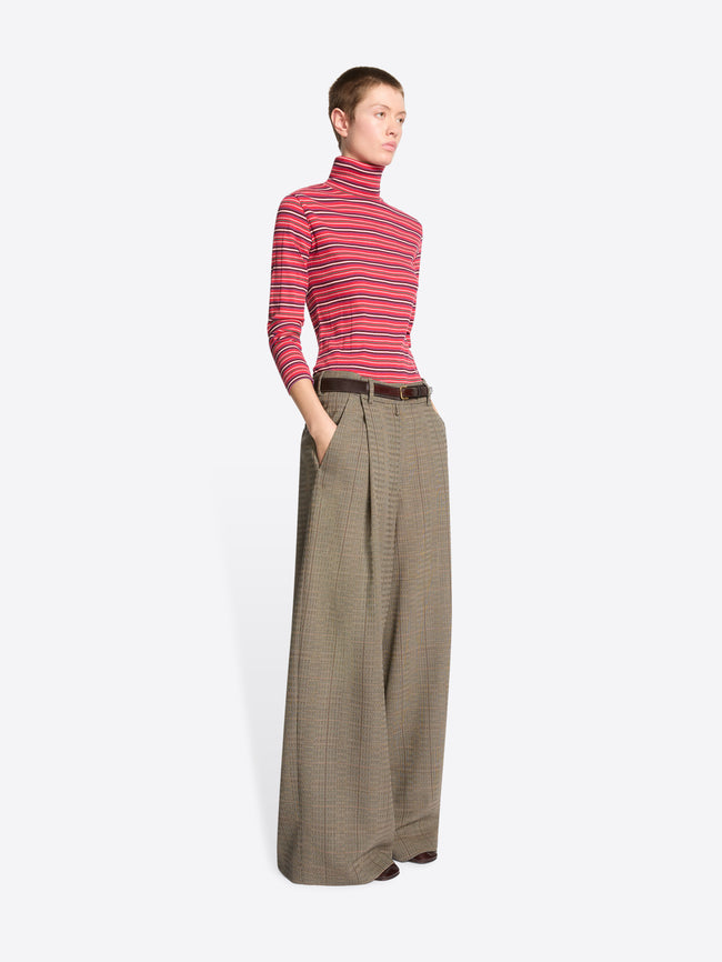A model stands against a light gray background wearing a red and pink striped turtleneck sweater paired with wide-leg brown trousers.