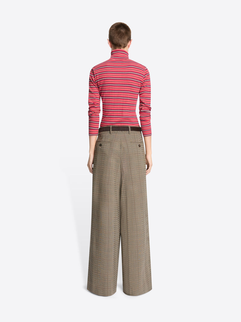 A model stands against a light gray background wearing a red and pink striped turtleneck sweater paired with wide-leg brown trousers.