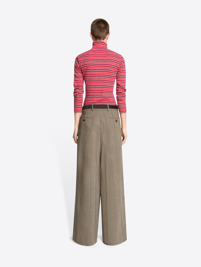 A model stands against a light gray background wearing a red and pink striped turtleneck sweater paired with wide-leg brown trousers.