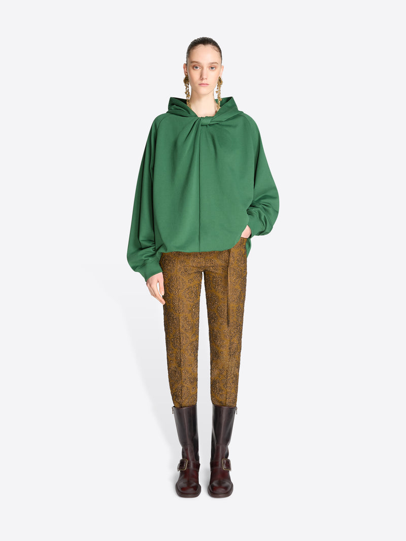 A model stands against a light gray background wearing a green oversized draped hoodie, brown patterned trousers, and dark brown leather boots.