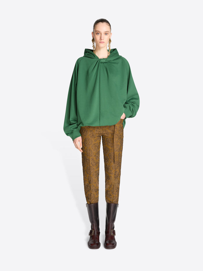 A model stands against a light gray background wearing a green oversized draped hoodie, brown patterned trousers, and dark brown leather boots.