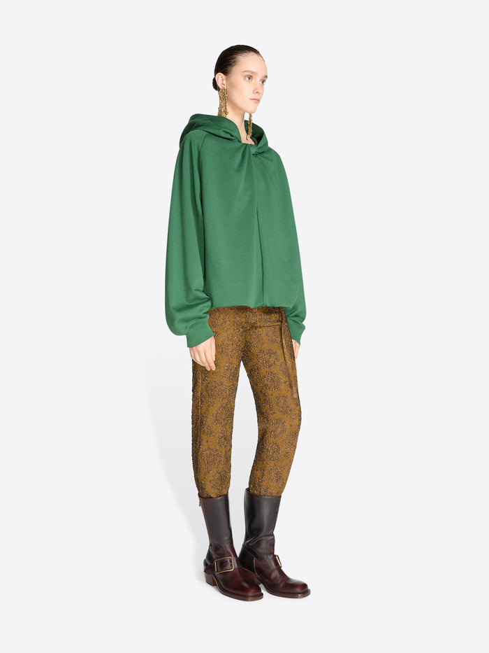 A model stands against a light gray background wearing a green oversized draped hoodie, brown patterned trousers, and dark brown leather boots.