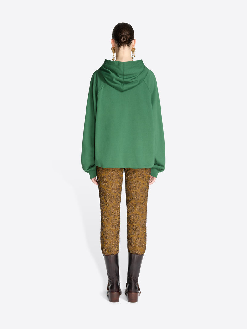 Back view of a model standing against a light gray background wearing a green oversized draped hoodie, brown patterned trousers, and dark brown leather boots.