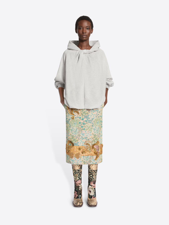 A model stands against a light gray background wearing a light gray oversized hoodie, a knee-length floral print skirt  and patterned tights with heels.