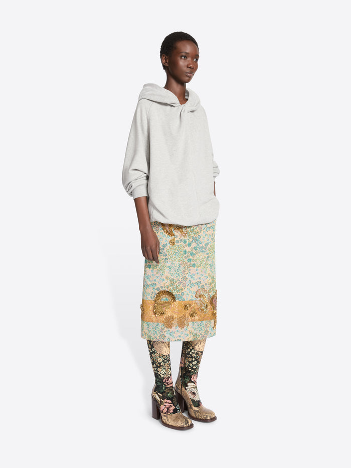 A model stands against a light gray background wearing a light gray oversized hoodie, a knee-length floral print skirt  and patterned tights with heels.