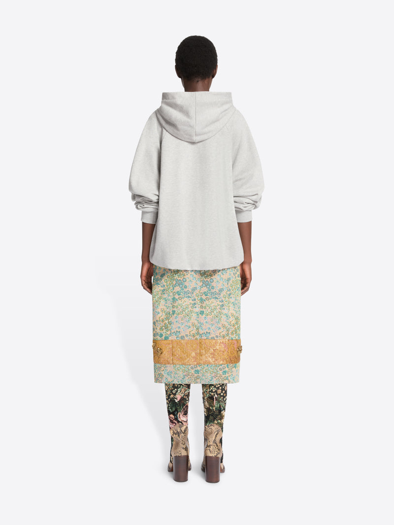 Back view of a model standing against a light gray background wearing a light gray oversized hoodie, a knee-length floral print skirt  and patterned tights with heels.