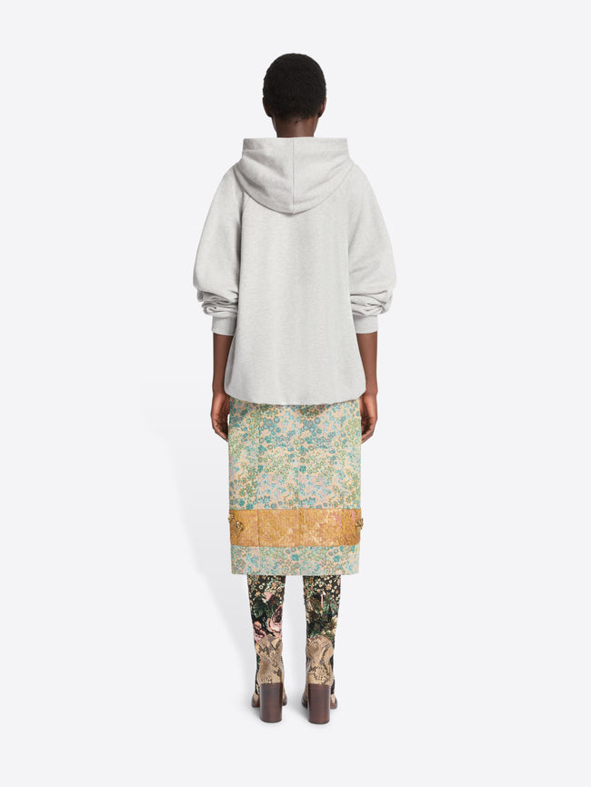 Back view of a model standing against a light gray background wearing a light gray oversized hoodie, a knee-length floral print skirt  and patterned tights with heels.