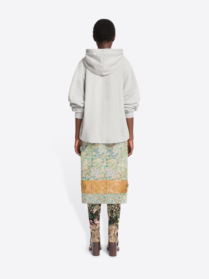 Back view of a model standing against a light gray background wearing a light gray oversized hoodie, a knee-length floral print skirt  and patterned tights with heels.