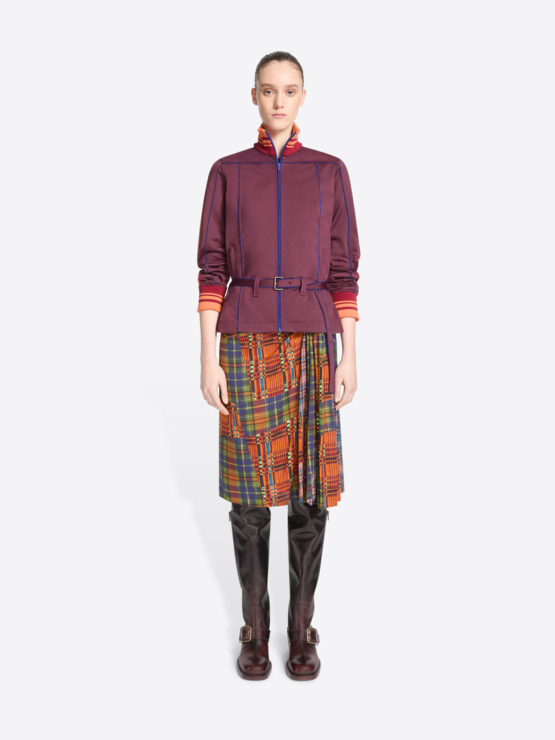 A model stands against a light gray background wearing a dark purple jacket with a belted waist, a multicolor plaid skirt and leather boots