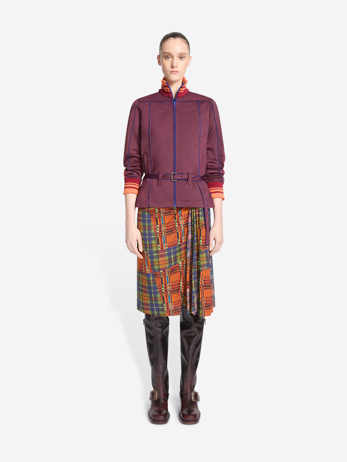 A model stands against a light gray background wearing a dark purple jacket with a belted waist, a multicolor plaid skirt and leather boots