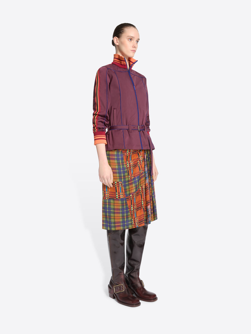 A model stands against a light gray background wearing a dark purple jacket with a belted waist, a multicolor plaid skirt and leather boots