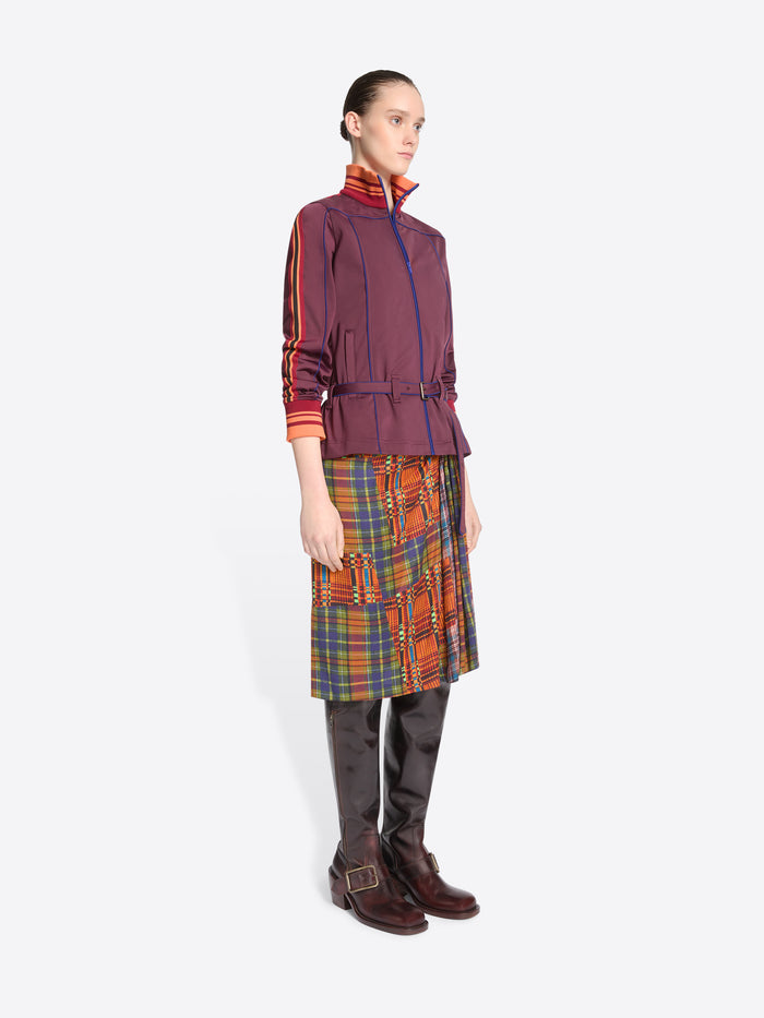 A model stands against a light gray background wearing a dark purple jacket with a belted waist, a multicolor plaid skirt and leather boots