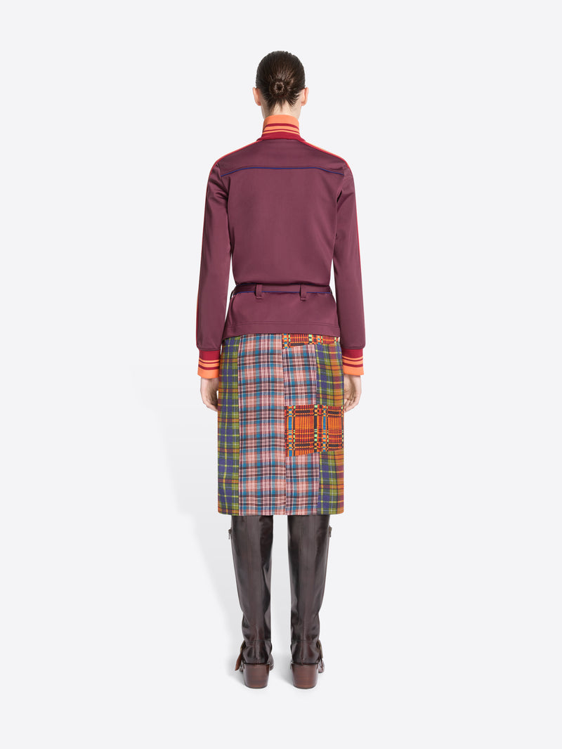 Back view of a model standing against a light gray background wearing a dark purple jacket with a belted waist, a multicolor plaid skirt and leather boots
