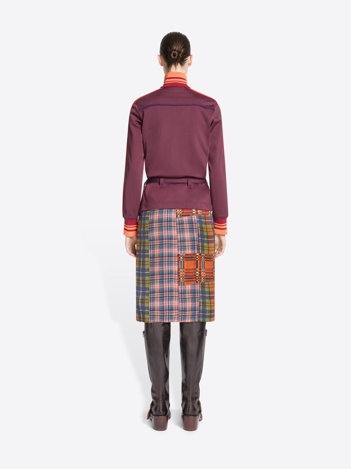 Back view of a model standing against a light gray background wearing a dark purple jacket with a belted waist, a multicolor plaid skirt and leather boots