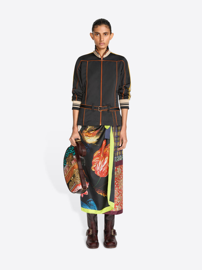 A model stands against a light gray background wearing a black long-sleeve jacket with yellow, orange and cream stripes, a matching black belt, and a long multicolor patterned skirt with a bright yellow-green border, holding a matching patterned handbag.