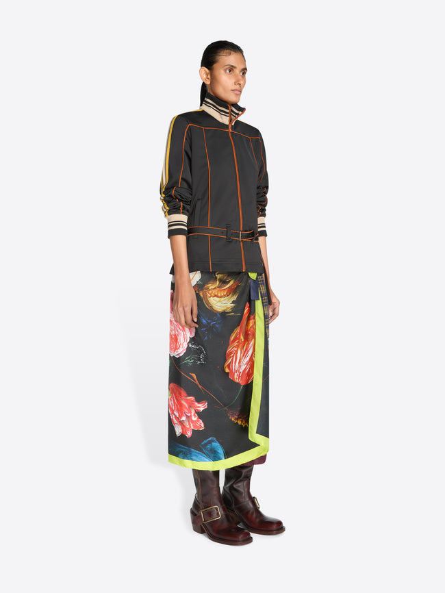 A model stands against a light gray background wearing a black long-sleeve jacket with yellow, orange and cream stripes, a matching black belt, and a long multicolor patterned skirt with a bright yellow-green border.