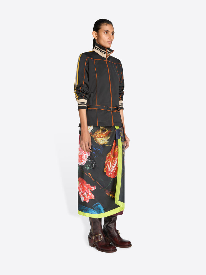 A model stands against a light gray background wearing a black long-sleeve jacket with yellow, orange and cream stripes, a matching black belt, and a long multicolor patterned skirt with a bright yellow-green border.