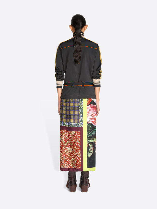 Back view of a model standing against a light gray background wearing a black long-sleeve jacket with yellow, orange and cream stripes, a matching black belt, and a long multicolor patterned skirt with a bright yellow-green border,  holding a matching patterned handbag.