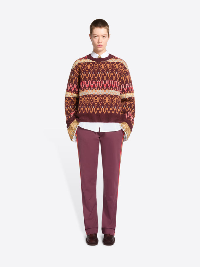A model stands against a light gray background wearing a patterned knit sweater, a white collared shirt underneath, mauve pants, and dark shoes.