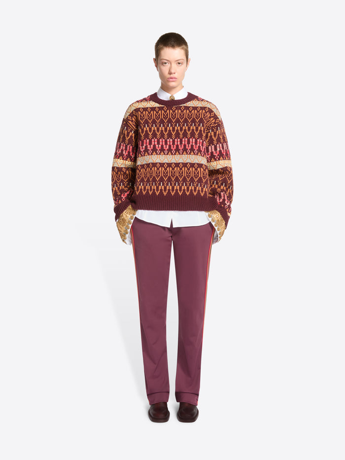 A model stands against a light gray background wearing a patterned knit sweater, a white collared shirt underneath, mauve pants, and dark shoes.