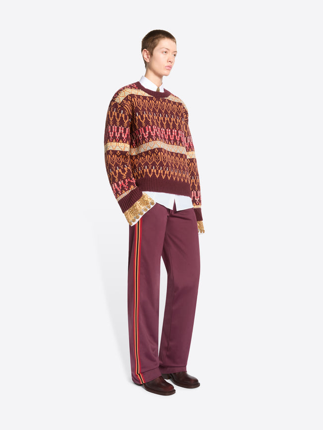 A model stands against a light gray background wearing a patterned knit sweater, a white collared shirt underneath, mauve pants, and dark shoes.