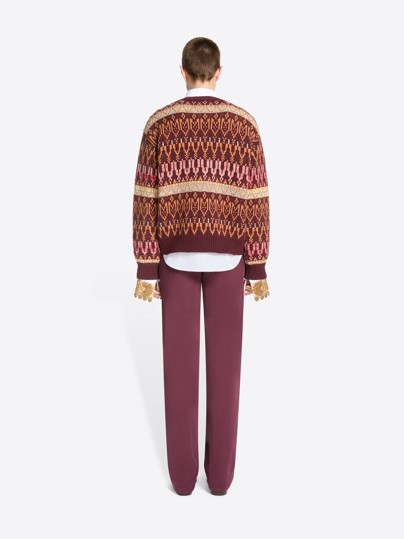 Back view of a model standing against a light gray background wearing a patterned knit sweater, a white collared shirt underneath, mauve pants, and dark shoes.