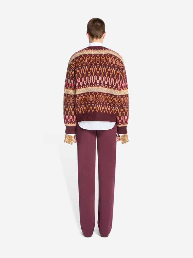 Back view of a model standing against a light gray background wearing a patterned knit sweater, a white collared shirt underneath, mauve pants, and dark shoes.