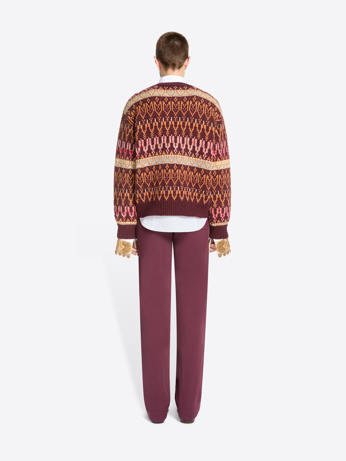 Back view of a model standing against a light gray background wearing a patterned knit sweater, a white collared shirt underneath, mauve pants, and dark shoes.