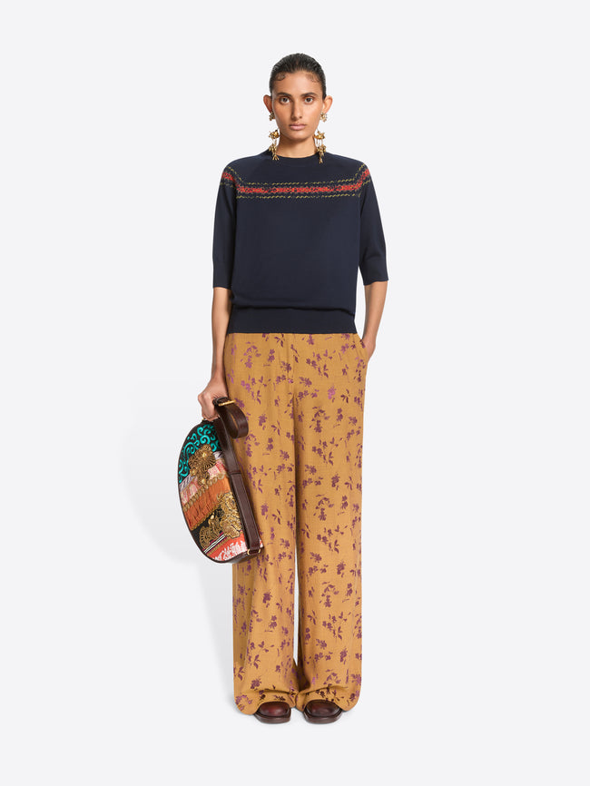 A model stands against a light gray background wearing a navy knit sweater with a colorful embroidered horizontal stripe across the chest, wide-leg orange patterned pants, and holding a colorful oval bag.