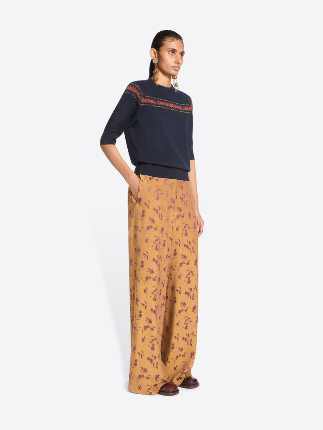 A model stands against a light gray background wearing a navy knit sweater with a colorful embroidered horizontal stripe across the chest, wide-leg orange patterned pants.
