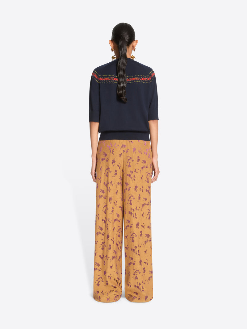 Back view of a model standing against a light gray background wearing a navy knit sweater with a colorful embroidered horizontal stripe across the chest, wide-leg orange patterned pants.