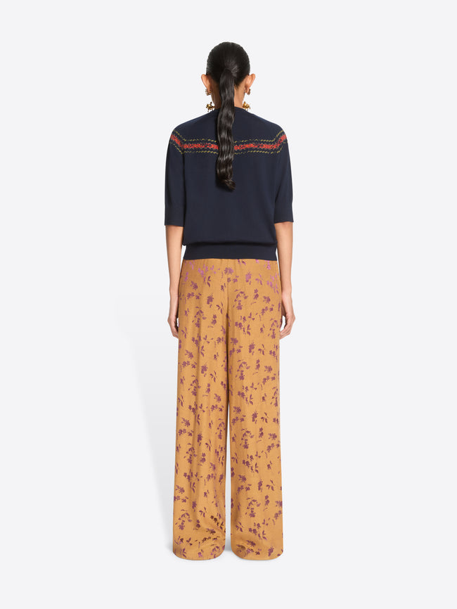 Back view of a model standing against a light gray background wearing a navy knit sweater with a colorful embroidered horizontal stripe across the chest, wide-leg orange patterned pants.