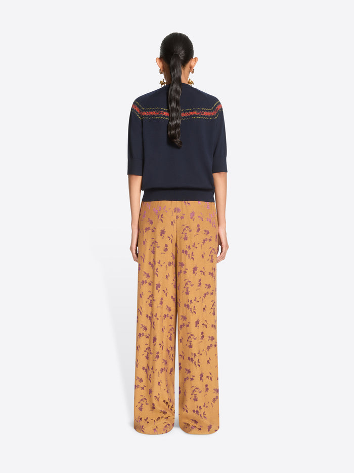 Back view of a model standing against a light gray background wearing a navy knit sweater with a colorful embroidered horizontal stripe across the chest, wide-leg orange patterned pants.