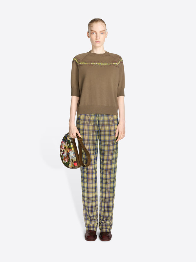 A model stands against a light gray background wearing a brown knit sweater with a colorful geometric pattern stripe across the chest and shoulders, olive green and yellow plaid trousers, and dark brown shoes, while holding a round floral-patterned handbag.