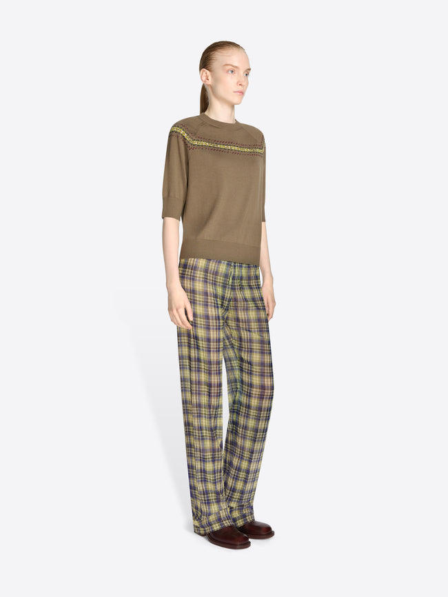A model stands against a light gray background wearing a brown knit sweater with a colorful geometric pattern stripe across the chest and shoulders, olive green and yellow plaid trousers, and dark brown shoes.